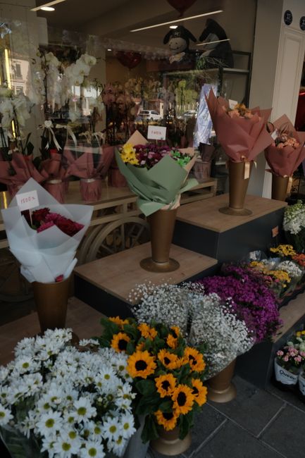 A display of fresh floral bouquets arranged in tall, cone-shaped, brown paper wrapping with decorative pink and beige tissue paper, positioned on a wooden table inside a flower shop. The bouquets feat
