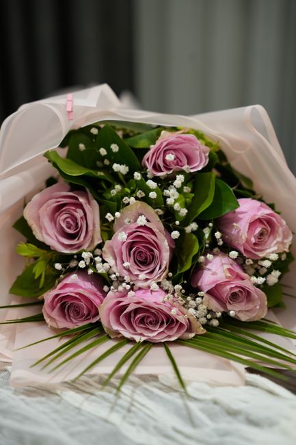 A bouquet of nine pale pink roses with tightly furled petals and a velvety texture, arranged with lush green leaves and delicate white baby's breath flowers. The flowers are wrapped in white tissue pa