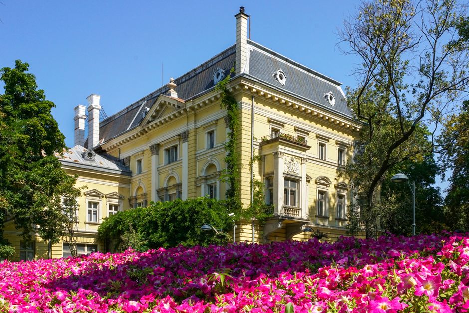 A historic, yellow-painted Victorian-style building with detailed architectural features, including decorative window frames and a slate roof, surrounded by lush green trees. In the foreground, there