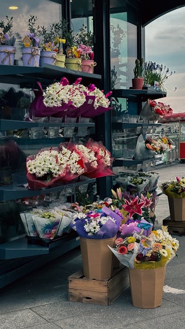 A display of fresh floral bouquets and potted plants arranged on black metal shelves and wooden crates outside a floral shop, featuring a variety of flowers such as white chrysanthemums, pink roses, p